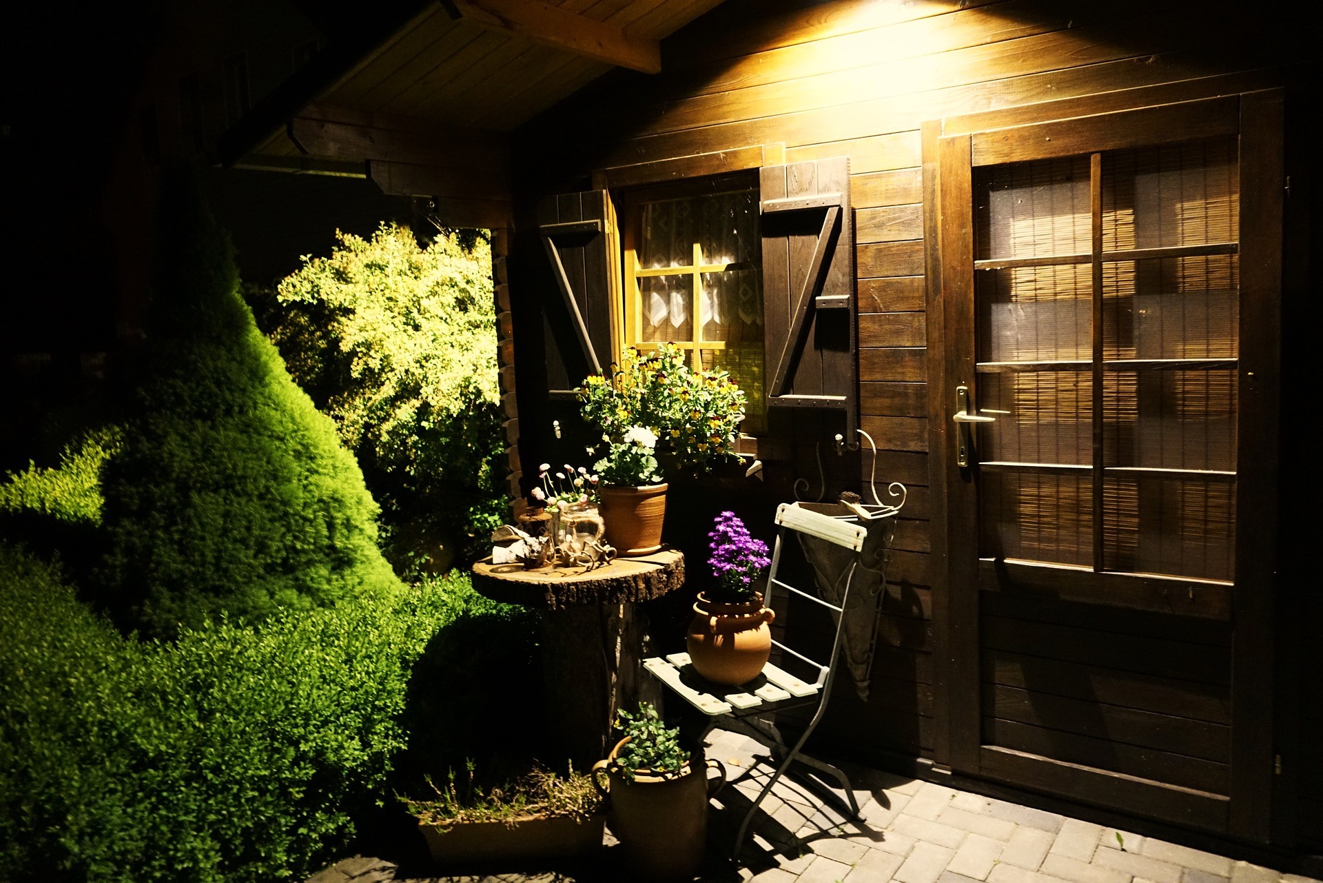 She Sheds at night with a yellow glowing porch light over a garden table and chair with potted plants on top